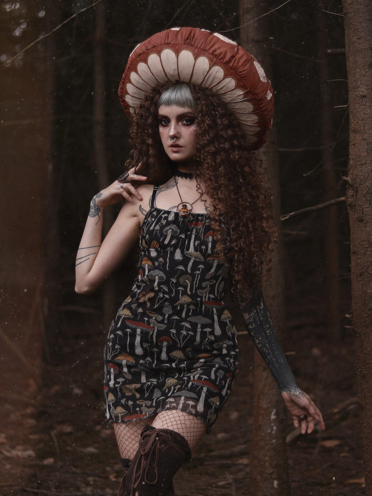 A girl with a mushroom hat stood in the woods, she is wearing a mushroom dress, fishnets & thigh high boots