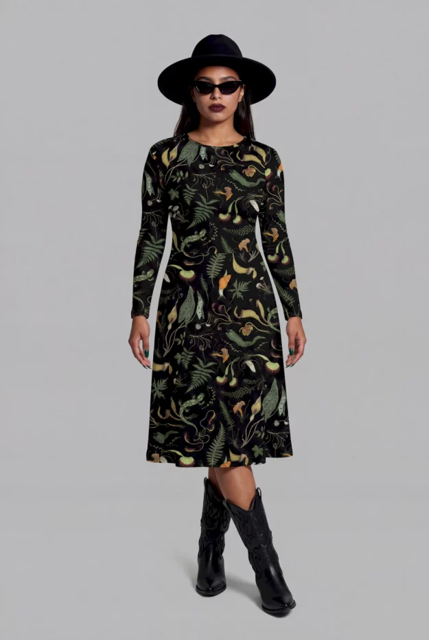 A woman wears the Cosmic Drifters Werewood Long Sleeve Midi Dress, a whimsigoth forest print piece, with a black hat, sunglasses, and black cowboy boots as she stands facing forward against a plain background.