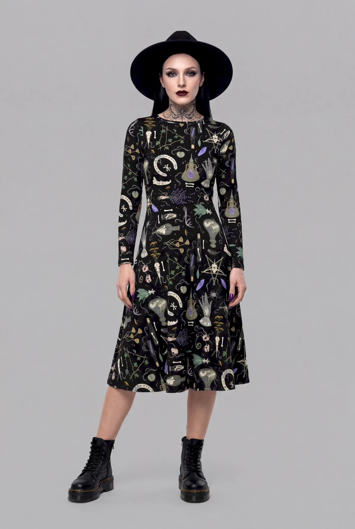 A model exudes a mystical aura in the Cosmic Drifters Witches' Altar Long Sleeve Midi Dress, styled with a black hat, dark make-up, and black boots against a plain grey background.