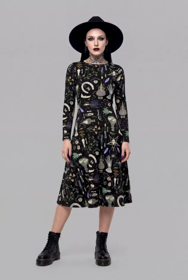 A model exudes a mystical aura in the Cosmic Drifters Witches' Altar Long Sleeve Midi Dress, styled with a black hat, dark make-up, and black boots against a plain grey background.