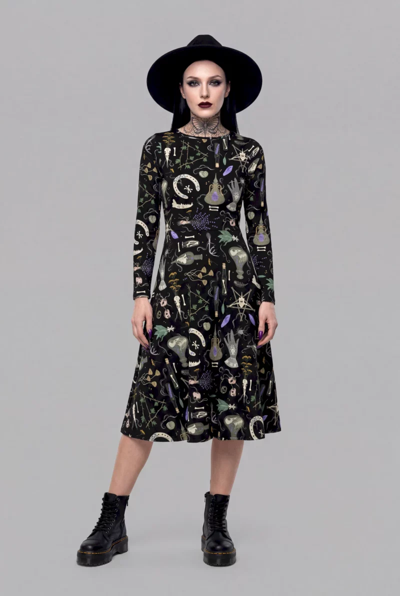 A model exudes a mystical aura in the Cosmic Drifters Witches' Altar Long Sleeve Midi Dress, styled with a black hat, dark make-up, and black boots against a plain grey background.