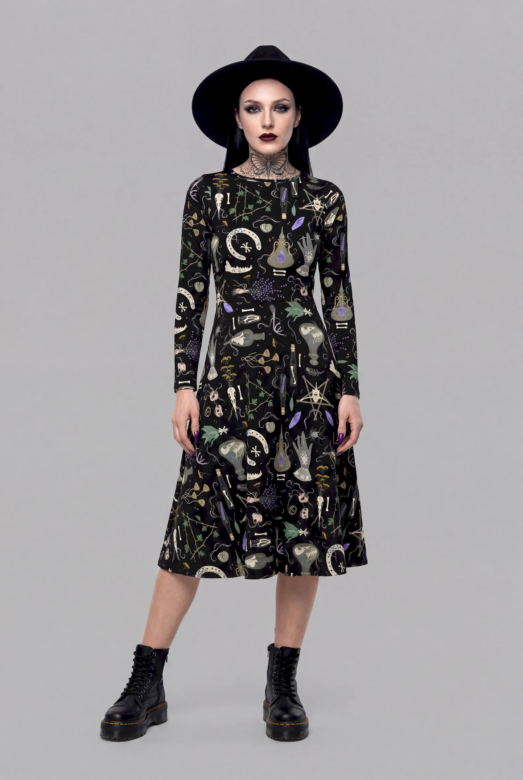 A model exudes a mystical aura in the Cosmic Drifters Witches' Altar Long Sleeve Midi Dress, styled with a black hat, dark make-up, and black boots against a plain grey background.