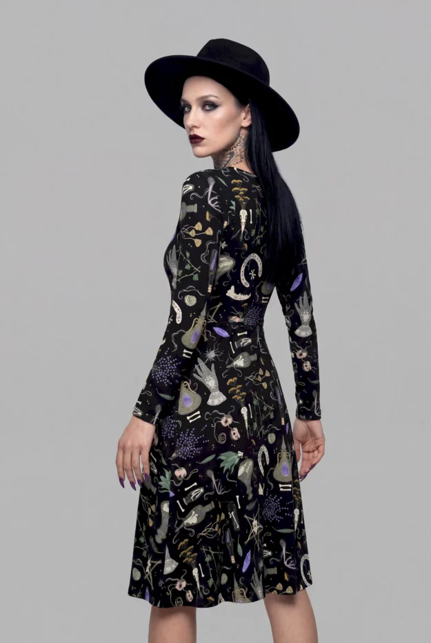 A woman in the Cosmic Drifters’ Witches Altar Long Sleeve Midi Dress and a black wide-brim hat stands sideways against a plain grey background, radiating a mystical aura.