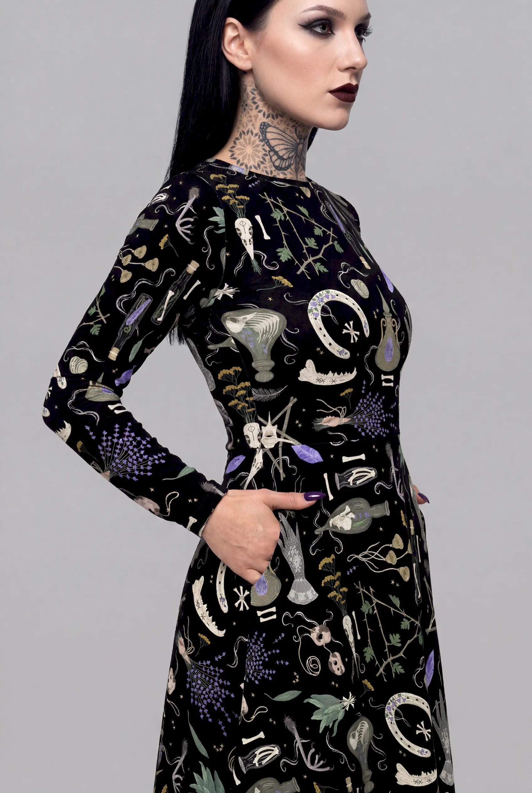 A dark-haired woman with a tattooed neck stands in profile, hands in pockets, wearing the Cosmic Drifters Witches Altar Long Sleeve Midi Dress adorned with mystical scientific and botanical illustrations.